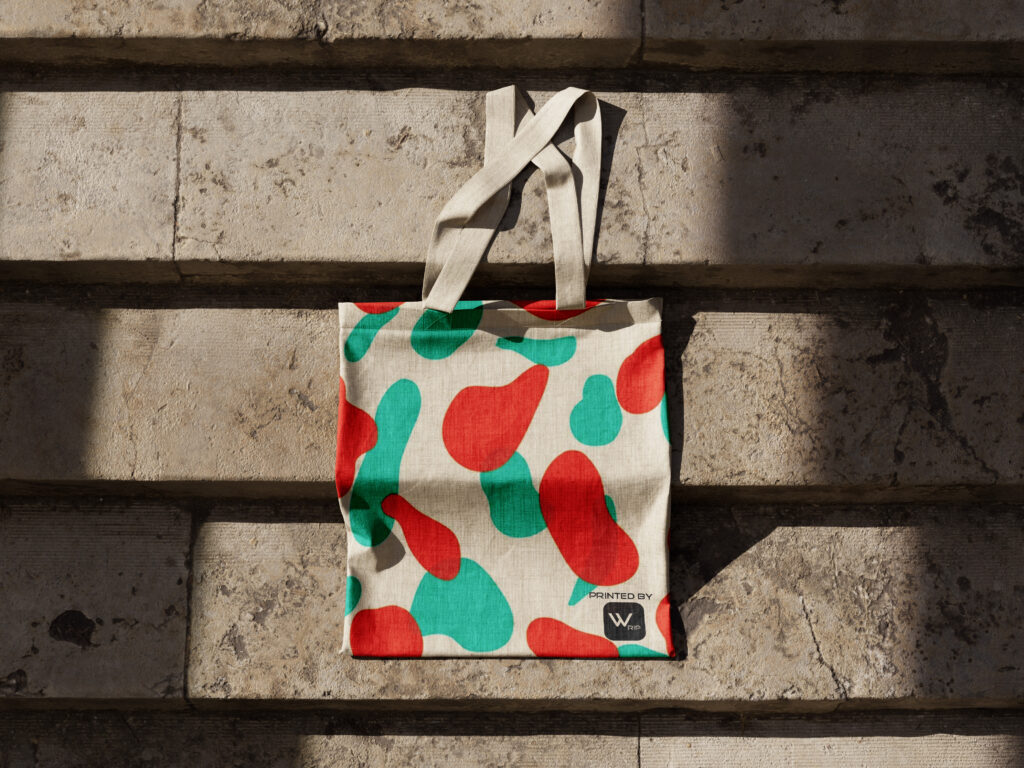 A stylish tote bag with a bold, abstract red and green print rests against an aged stone wall, bathed in the soft light of the sun, highlighting the natural and eco-friendly vibe of the bag