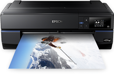 This image showcases an Epson flatbed printer, likely a model designed for high-quality, large-format printing. The printer is predominantly black with a sleek and contemporary design, featuring a control panel with buttons and a small display screen on the front right side. A print is emerging from the printer, illustrating a vibrant and detailed image of a mountaineer ascending a snowy peak, which is a testament to the printer's ability to produce vivid color output. The image of the mountaineer against the stark white of the snow emphasizes the printer's capability to handle complex and colorful images. The Epson logo is prominently displayed on the top center of the printer, indicating the brand. The overall setting suggests a professional or semi-professional environment where high-quality prints are produced.