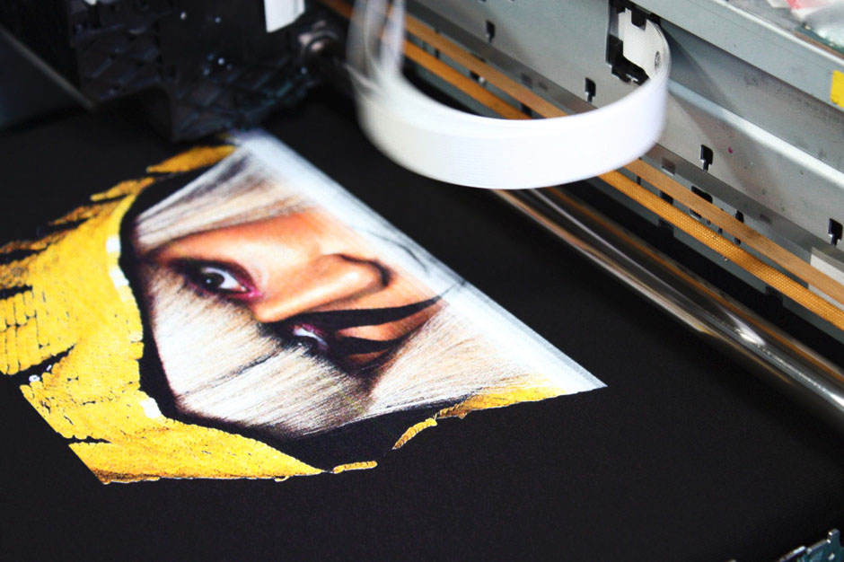 This image captures a close-up of a direct-to-garment (DTG) printing process in action. We can see a portion of a T-shirt being printed with an intricate design featuring what appears to be an eye, surrounded by splashes of yellow paint against a black fabric background. The print is vivid, displaying fine details and a wide range of colors that suggest high-quality printing technology. Part of the printer's carriage is visible in the upper portion of the image, with a white belt that likely moves the garment through the machine for printing. The metal components and industrial design of the printer indicate that it's a professional-grade machine designed for producing detailed, durable prints on fabric. The image demonstrates the machine's capability to create complex, color-rich images directly onto clothing.